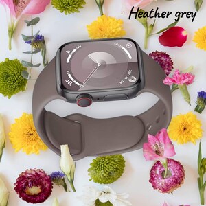 Op de afbeelding: Een heather grey smartwatch met een bijpassende band, met de tijd op een zwart scherm. Het horloge heeft een rode knop en is omringd door kleurrijke bloemen, waaronder roze, gele en witte bloemen.