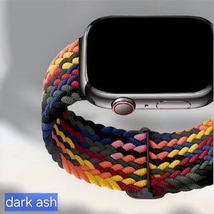 Puede incluir: Un reloj inteligente gris oscuro con pantalla negra y una correa trenzada multicolor. La correa presenta una mezcla de colores como amarillo, naranja, rojo, verde y azul. El texto "dark ash" es visible en la parte inferior.