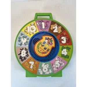 May include: A vintage green and blue See 'N Say toy with a handle. The toy features a rotating dial with numbers 1-10, each accompanied by colorful illustrations of animals and objects. The center has a dial that says "Count with Colors".