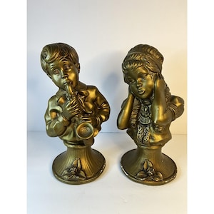 Puede incluir: Dos bustos dorados de niños. Uno toca el saxofón, el otro se tapa los oídos. Ambos están sobre pedestales con detalles florales. Las estatuas tienen una estética vintage y probablemente sean objetos decorativos.