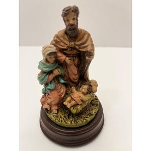 May include: A religious figurine depicting the Nativity scene. The statue features Mary, Joseph, and baby Jesus in a manger. The figures are dressed in robes of various colors, including peach, green, and brown. The base is a dark brown color.