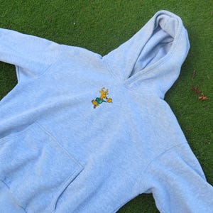May include: Light blue hoodie with a kangaroo pocket and a drawstring hood. The hoodie features an embroidered cartoon deer wearing a green outfit and riding a skateboard. The hoodie is made of a soft, comfortable material.