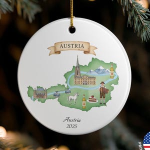 May include: White ceramic Christmas ornament featuring a map of Austria with illustrations of landmarks and a traditional figure. The word "Austria" is displayed on a banner, with "Austria 2025" below. The ornament is hanging from a gold string.
