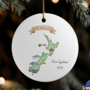 May include: A white ceramic Christmas ornament with a gold string, featuring a map of New Zealand. The ornament has a banner that reads "NEW ZEALAND" and the text "New Zealand 2025". The ornament is hanging on a Christmas tree.