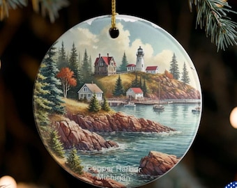 May include: Circular ceramic Christmas ornament depicting a coastal scene with a lighthouse, houses, and boats. It has a gold hanging string. The text "Copper Harbor Michigan" is printed on the bottom of the ornament.