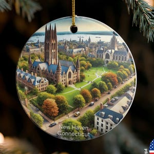 May include: Round Christmas ornament featuring a detailed aerial view of New Haven, Connecticut, with a prominent church and surrounding buildings. The ornament has a gold string for hanging and the text "New Haven Connecticut".