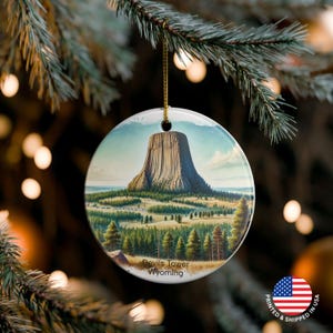 Puede incluir: Un adorno navideño redondo con una ilustración del Devils Tower en Wyoming. El adorno tiene una cuerda dorada para colgar y el texto "Devils Tower Wyoming". El fondo está borroso con luces.