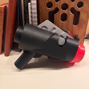 May include: A black and red 3D-printed toy gun with a handle and a red tip. The toy gun is resting on a white surface. A gray rectangular piece is behind the toy gun.