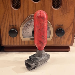 May include: A red and gray bottle stopper with a cylindrical top and a rectangular base. The stopper is placed in front of a vintage wooden radio.