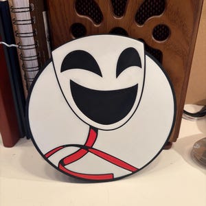 May include: A white, circular coaster with a smiling face design. The face features a large black mouth and eyes, with red accents. The coaster is placed on a white surface, with a wooden radio and notebooks in the background.