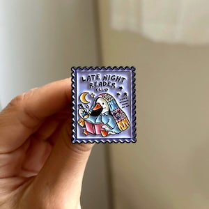 May include: A rectangular enamel pin with a purple border featuring a cartoon goose reading a book. The pin reads "Late Night Reader Club" with a crescent moon and stars. The goose wears a nightcap and scarf.