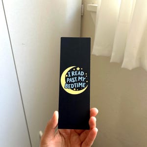 May include: A black bookmark with the text "I READ PAST MY BEDTIME" in white and blue, with a yellow crescent moon and stars design. The bookmark is rectangular and held in a hand.