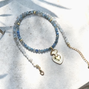 Könnte beinhalten: Eine Perlenkette mit kleinen, facettierten blauen und durchscheinend grauen Steinen in einem Farbverlauf. Die Kette enthält kleine goldfarbene Metallabstandshalter und einen zentralen goldfarbenen herzförmigen Anhänger mit gewelltem Rand.