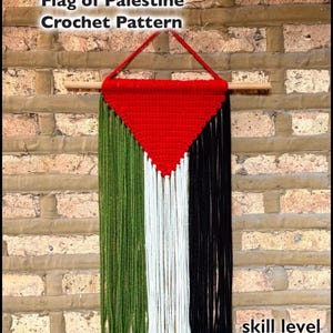 May include: A crochet pattern depicting the Flag of Palestine, suspended from a wooden dowel. The design includes a red triangle, and green, white, and black vertical stripes. The text "skill level beginner" is visible.