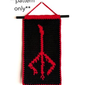 May include: A black crochet banner with a red border and a red symbol in the center. The banner is suspended from a black rod with a red yarn loop at the top. The text "pattern only" is at the top.