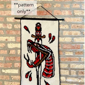 Könnte beinhalten: Gehäkeltes Wandbehang mit einem Schlangen- und Dolch-Design in Rot, Schwarz und Gold auf cremefarbenem Hintergrund. Das Stück hat einen schwarzen Rand und den Text "pattern only".
