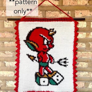 May include: Crocheted wall hanging featuring a red devil character with a trident, standing on a dice. The artwork is framed with a red scalloped border and a white background. The text "pattern only" is visible.