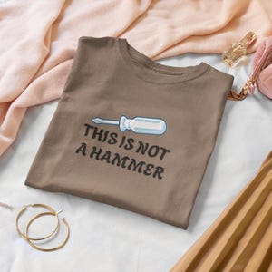 May include: A brown t-shirt featuring the text "THIS IS NOT A HAMMER" and a screwdriver graphic. The image includes a pink scarf, gold hoop earrings, a pink quilted cosmetic bag, and a pleated skirt.