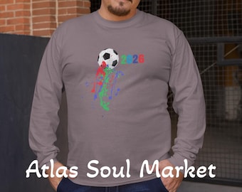 May include: A long-sleeved grey top featuring a soccer ball design with the numbers "2026" in red and blue. The shirt has colourful paint splatters. The words "Atlas Soul Market" are printed on the shirt and in a white box in the upper left corner.