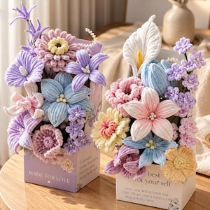 Könnte beinhalten: Zwei rechteckige Schachteln mit Arrangements aus gehäkelten Blumen in Pastelltönen wie Lila, Blau, Rosa und Gelb. Die linke Schachtel ist helllila mit dem Text MADE FOR LOVE, die rechte ist weiß und trägt unter anderem den Text best of your self.