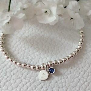 May include: Silver beaded bracelet featuring a heart-shaped charm with the letter "D" and a small blue gemstone. The bracelet is presented on a white textured surface, with white flowers in the background.