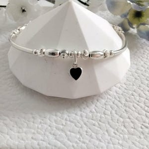 May include: A silver-toned bangle bracelet featuring a series of smooth and textured metallic beads along the band. A small, solid black heart-shaped charm hangs from the centre of the bracelet, creating a distinct contrast against the metallic finish.