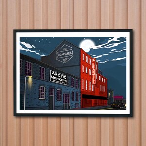 May include: A framed art print featuring a stylized illustration of a building at night. The building is divided into blue and red sections, with the text "Arctic Monkeys" and "Leadmill" visible. The night sky has a full moon.