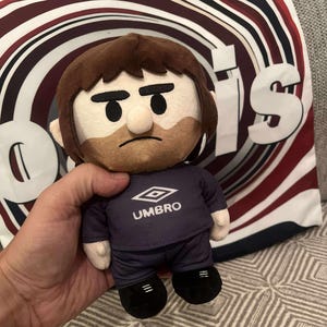 Puede incluir: Un muñeco de peluche con cabeza y barba marrones, que lleva un uniforme Umbro azul marino. El muñeco tiene una expresión seria. El fondo incluye una almohada con un diseño rojo, blanco y azul marino.