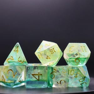 May include: Set of seven polyhedral dice in shades of green and yellow. The dice are transparent with gold-colored numbers. The dice are arranged on a dark surface, suitable for tabletop gaming.