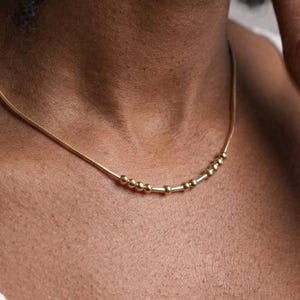 May include: A delicate gold necklace featuring a thin chain and small, evenly spaced beads. The necklace is worn around the neck and rests on a white top. The beads are clustered in groups.