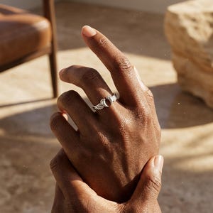 May include: A silver ring featuring a wing design on either side of a central, dark red stone. The ring is worn on a finger, showcasing its intricate details and craftsmanship. The ring's design is elegant and eye-catching.