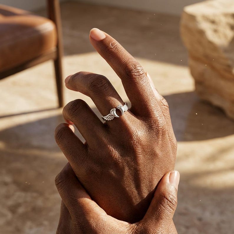 May include: A silver ring featuring a wing design on either side of a central, dark red stone. The ring is worn on a finger, showcasing its intricate details and craftsmanship. The ring's design is elegant and eye-catching.