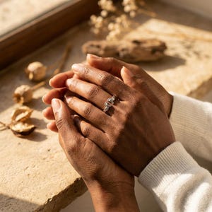 May include: A silver ring with a heart and wing design. The ring is worn on a finger, with the hands in the foreground. The ring is the focal point, with a neutral background. The ring is a piece of jewelry.