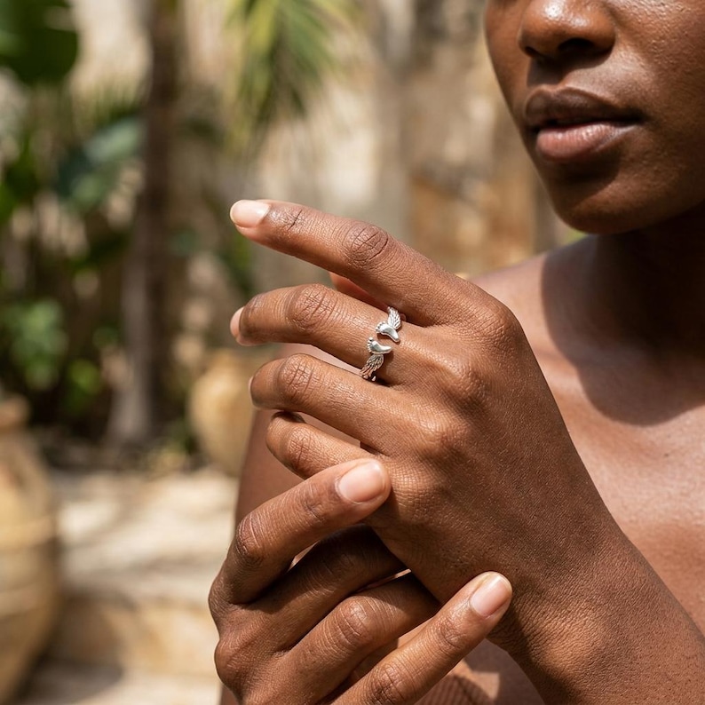 May include: A silver ring with a detailed design featuring two stylized bird wings. The ring is worn on a finger, showcasing its intricate craftsmanship and elegant aesthetic. The ring's design is visible against the skin.