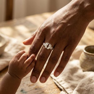 May include: A silver ring featuring two baby footprints with small wings on either side. The ring is on a person's finger, with a baby's hand reaching out to hold the finger. The background includes a wooden table and a neutral-colored cloth.