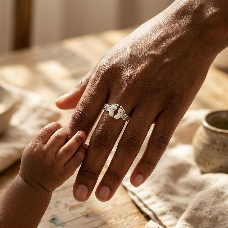 May include: A silver ring featuring two baby footprints with small wings on either side. The ring is on a person's finger, with a baby's hand reaching out to hold the finger. The background includes a wooden table and a neutral-colored cloth.