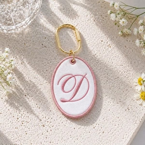 Puede incluir: Un llavero de forma ovalada con un centro de tela blanca bordado con una letra D rosa en cursiva. El llavero está enmarcado por un borde cosido en color rosa y unido a una anilla ovalada de metal dorado para una sujeción segura.