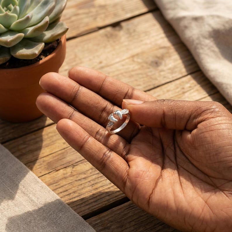 May include: A silver ring featuring a baby feet design. The ring is held in a hand, with the ring's details clearly visible. The ring is on a wooden surface, with a succulent plant in the background.