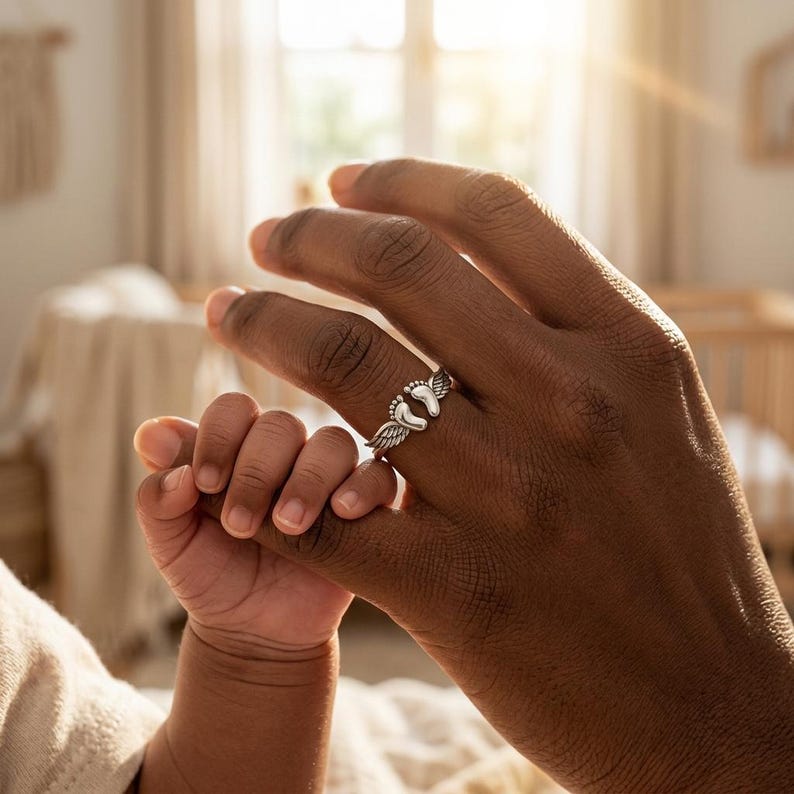 May include: A silver ring featuring two baby footprints with angel wings. The ring is worn on a finger, with a baby's hand holding the finger. The ring is a symbol of new life and family.