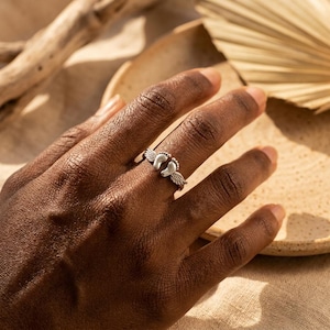 May include: A silver ring featuring a design of baby footprints with angel wings. The ring is worn on a hand, with a neutral background. The ring is a symbol of new beginnings and is a thoughtful gift for new parents.