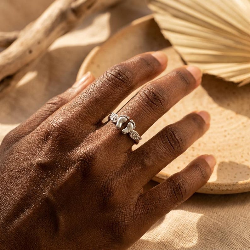 May include: A silver ring featuring a design of baby footprints with angel wings. The ring is worn on a hand, with a neutral background. The ring is a symbol of new beginnings and is a thoughtful gift for new parents.