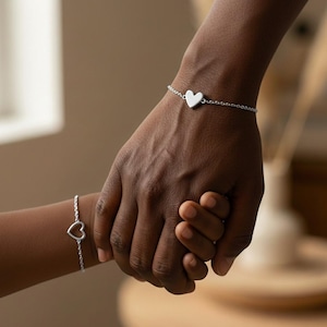 Op de afbeelding: Twee zilveren hartarmbanden zijn zichtbaar op twee polsen, een volwassen en een kind. De armband van de volwassene heeft een massieve hartbedel, terwijl die van het kind een open hartontwerp heeft. De armbanden zijn verbonden door delicate kettingen.