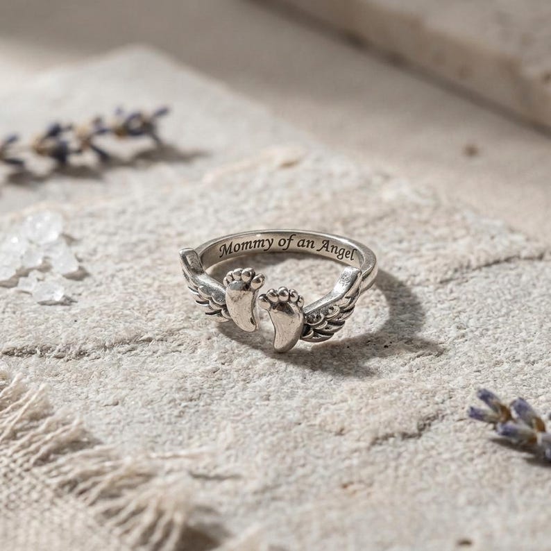 May include: A silver ring with the inscription "Mommy of an Angel." The ring features a pair of baby footprints and angel wings. The ring is set on a textured, light-colored surface.
