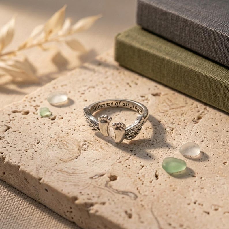 May include: A silver ring with the inscription "Mommy of an Angel." The ring features a pair of baby footprints and angel wings. The ring is displayed on a textured, light-colored surface, with decorative elements in the background.