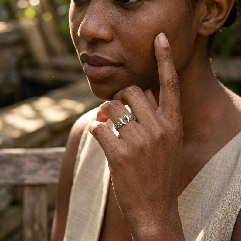 May include: A silver ring with a crescent moon design. The ring features a detailed, textured band and a prominent moon motif. The ring is worn on a finger, showcasing the intricate craftsmanship and design of the jewelry.