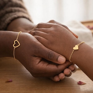 Może przedstawiać: Dwie złote bransoletki w kształcie serca są pokazane na ciemnych odcieniach skóry. Jedna bransoletka ma otwarte serce, a druga pełne serce. Bransoletki są na delikatnych łańcuszkach. Tło to drewniana powierzchnia.