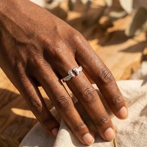 May include: A silver ring featuring a design of baby feet with angel wings. The ring is worn on a finger, showcasing the detailed craftsmanship. The ring is a symbol of love and protection.