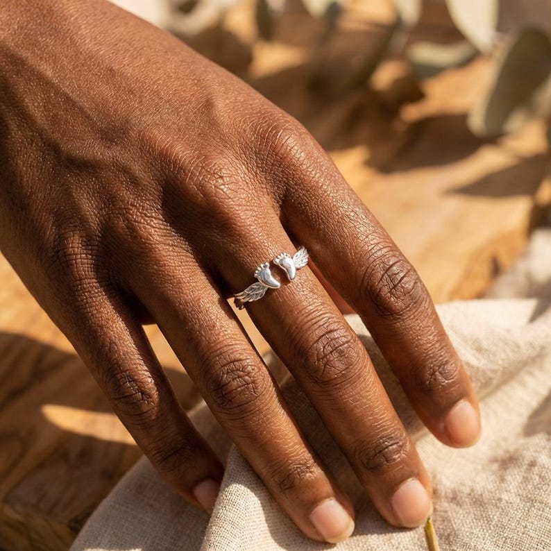 May include: A silver ring featuring a design of baby feet with angel wings. The ring is worn on a finger, showcasing the detailed craftsmanship. The ring is a symbol of love and protection.