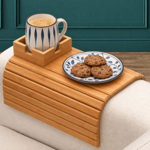 May include: A flexible bamboo armrest tray featuring a square holder for a white mug with blue vertical leaf patterns. A round white plate with blue leaf motifs holds three chocolate chip cookies, resting on the wooden slats of the tray.