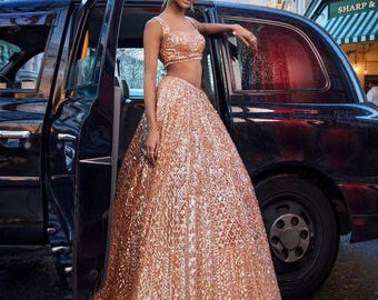 May include: A rose gold sequined two-piece formal gown. The top is a crop top with thin straps, and the skirt is a full, floor-length skirt. The outfit is worn in front of a black taxi cab.